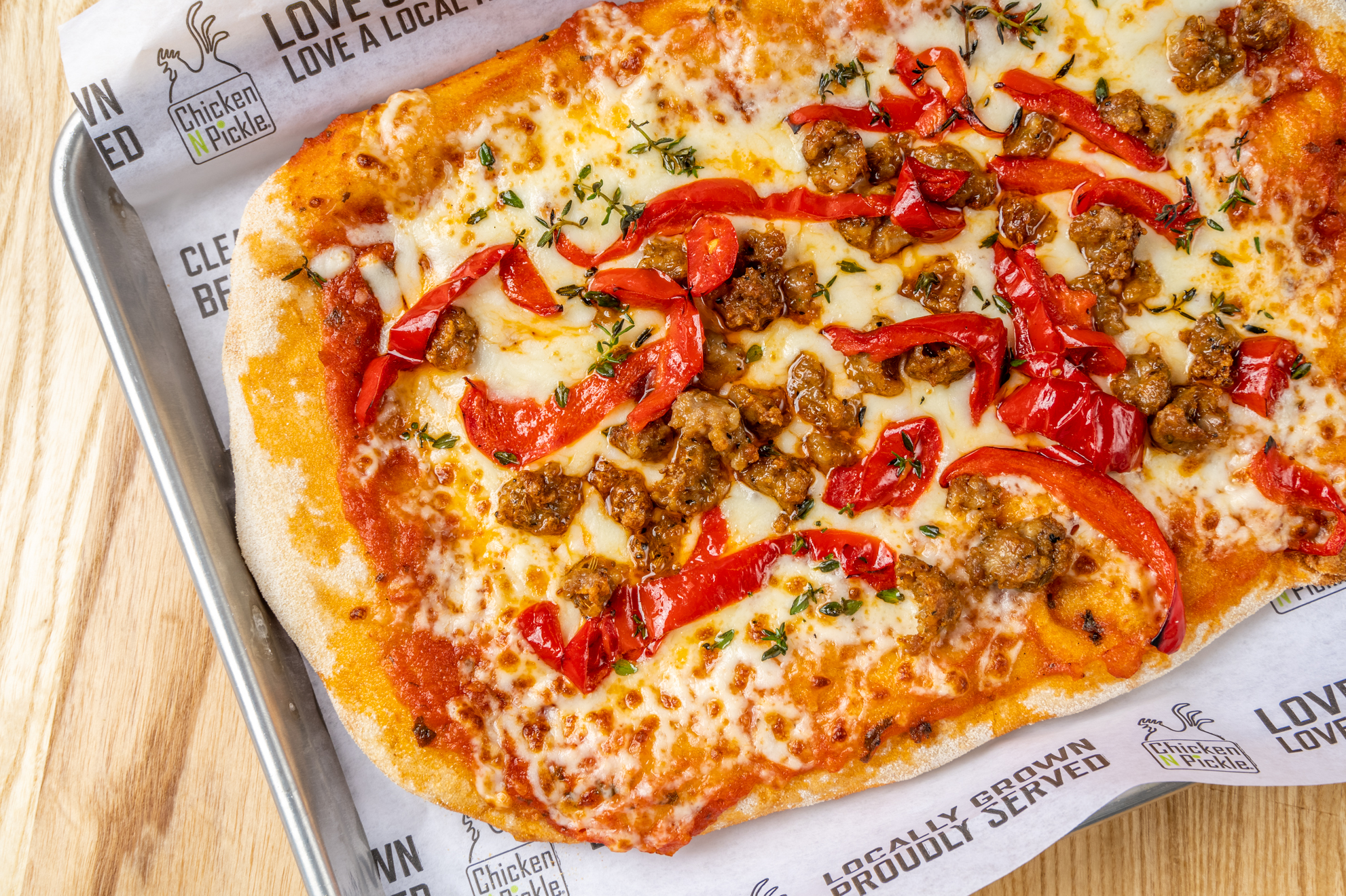 Rectangular pizza topped with melted cheese, sausage pieces, and sliced red bell peppers on a sheet pan lined with paper that has Chicken N Pickle branding. The crust is golden and slightly crispy.