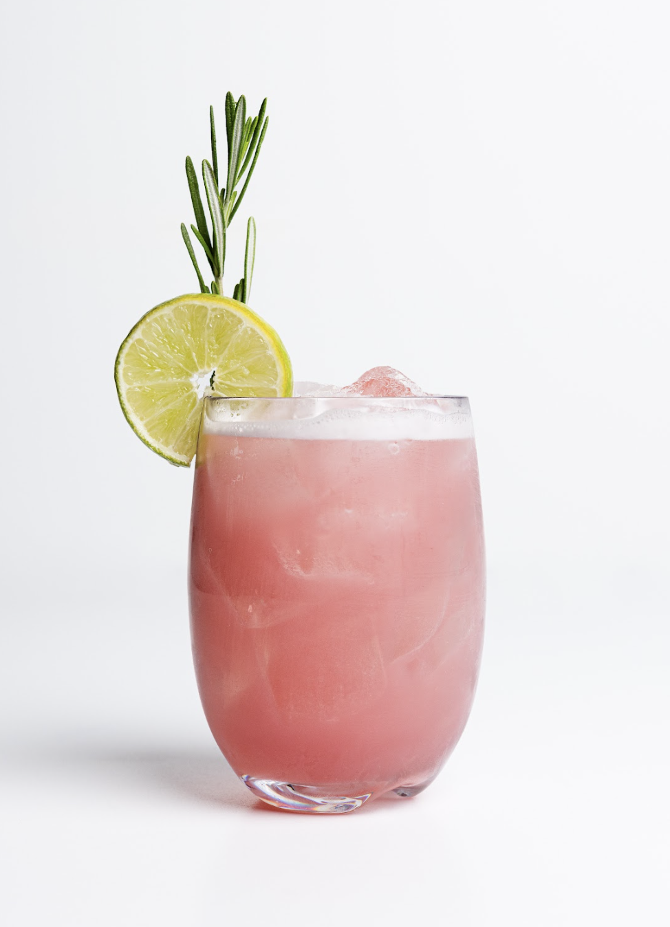 A pink iced drink from the Drink Menu sits in a glass, garnished with a slice of lime and a sprig of rosemary, set against a plain white background.