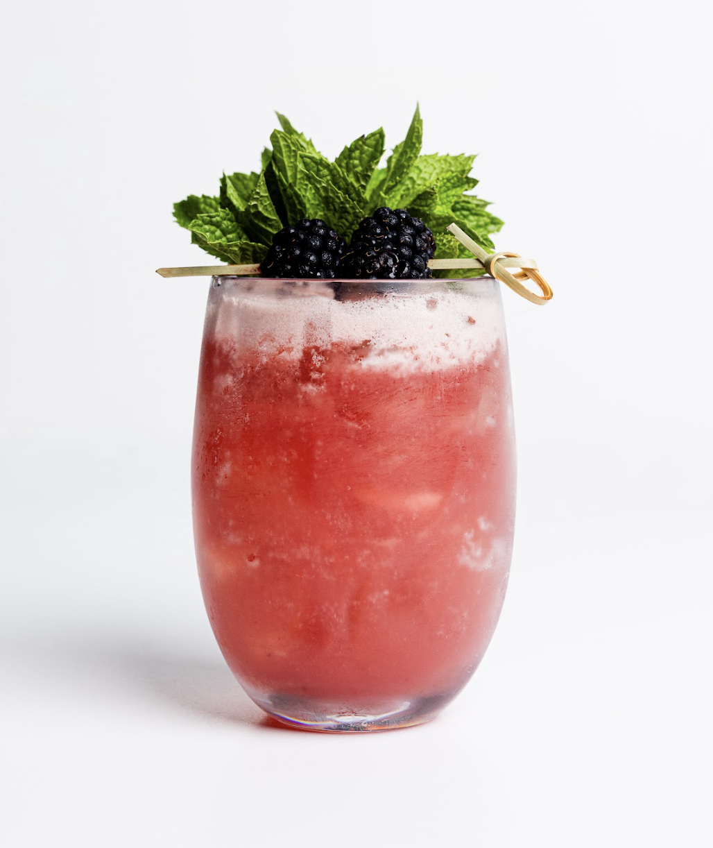 A vibrant pink cocktail from the Menu sits in a glass, topped with a skewer of blackberries and fresh mint leaves against a white background.