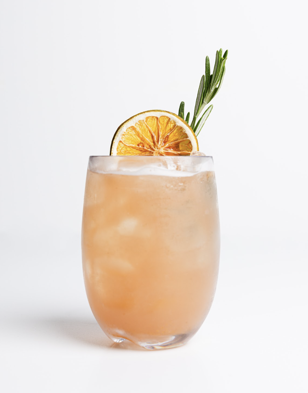 A pink-orange cocktail from the Menu sits in a clear glass filled with ice, garnished with a dried orange slice and a sprig of rosemary, set against a plain white background.