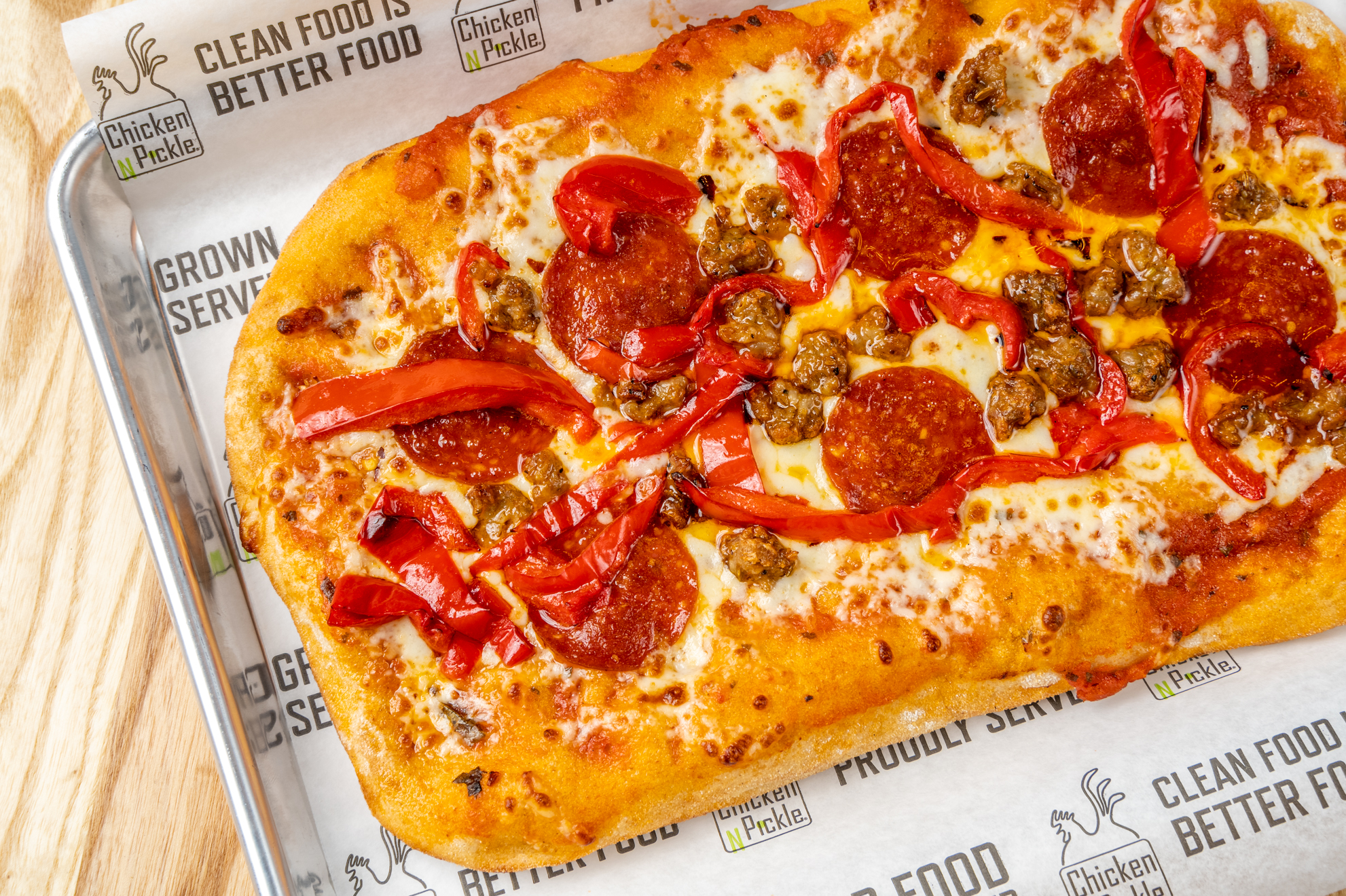 A rectangular flatbread pizza topped with melted cheese, pepperoni, sliced red peppers, and sausage pieces, served on branded parchment paper on a metal tray.