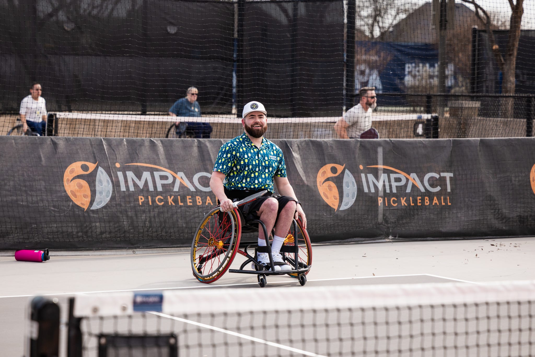 A man in a wheelchair, wearing a colorful shirt and white cap, smiles while playing pickleball on an outdoor court with IMPACT PICKLEBALL banners in the background.