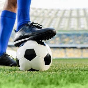 A soccer player wearing blue socks and black cleats stands on a grass field with one foot resting on a black and white soccer ball inside a stadium.