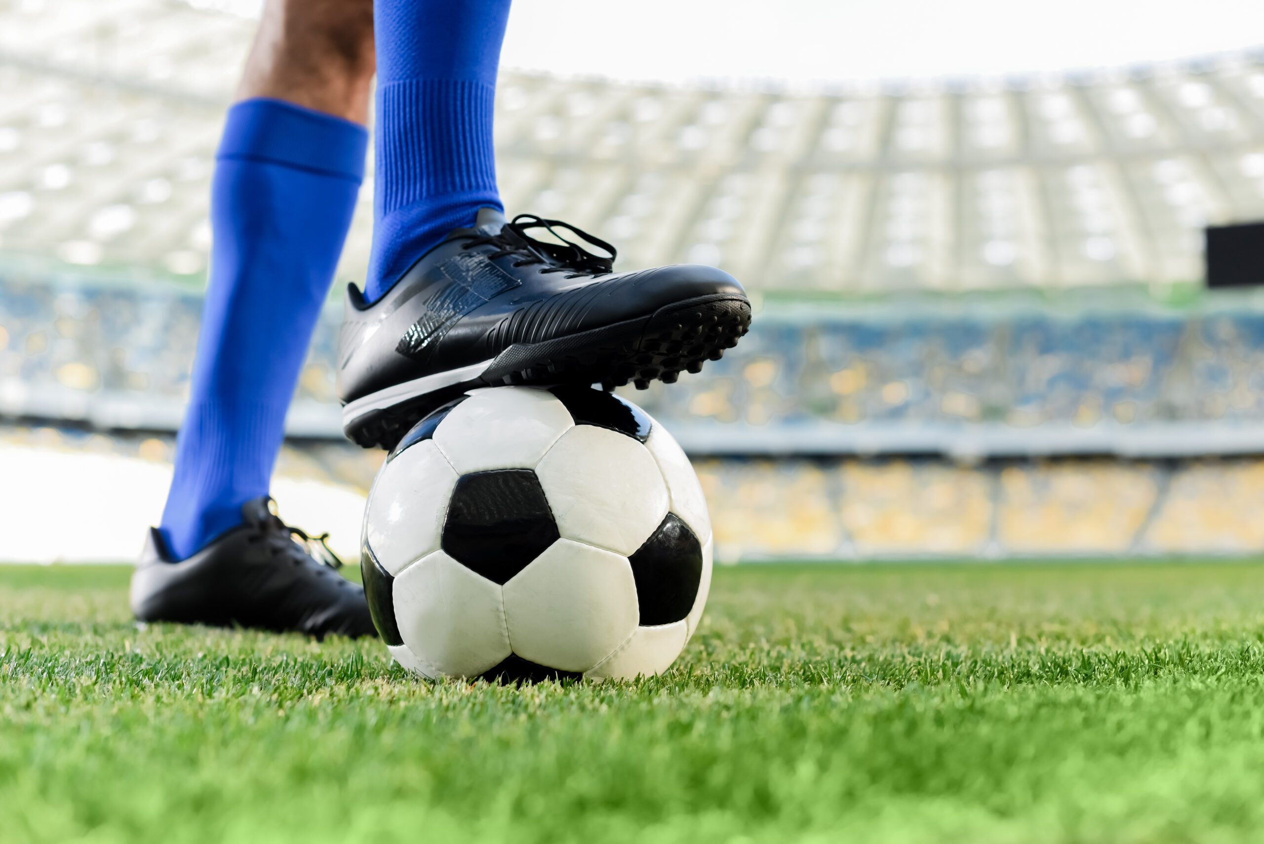 A soccer player wearing blue socks and black cleats stands on a grass field with one foot resting on a black and white soccer ball inside a stadium.