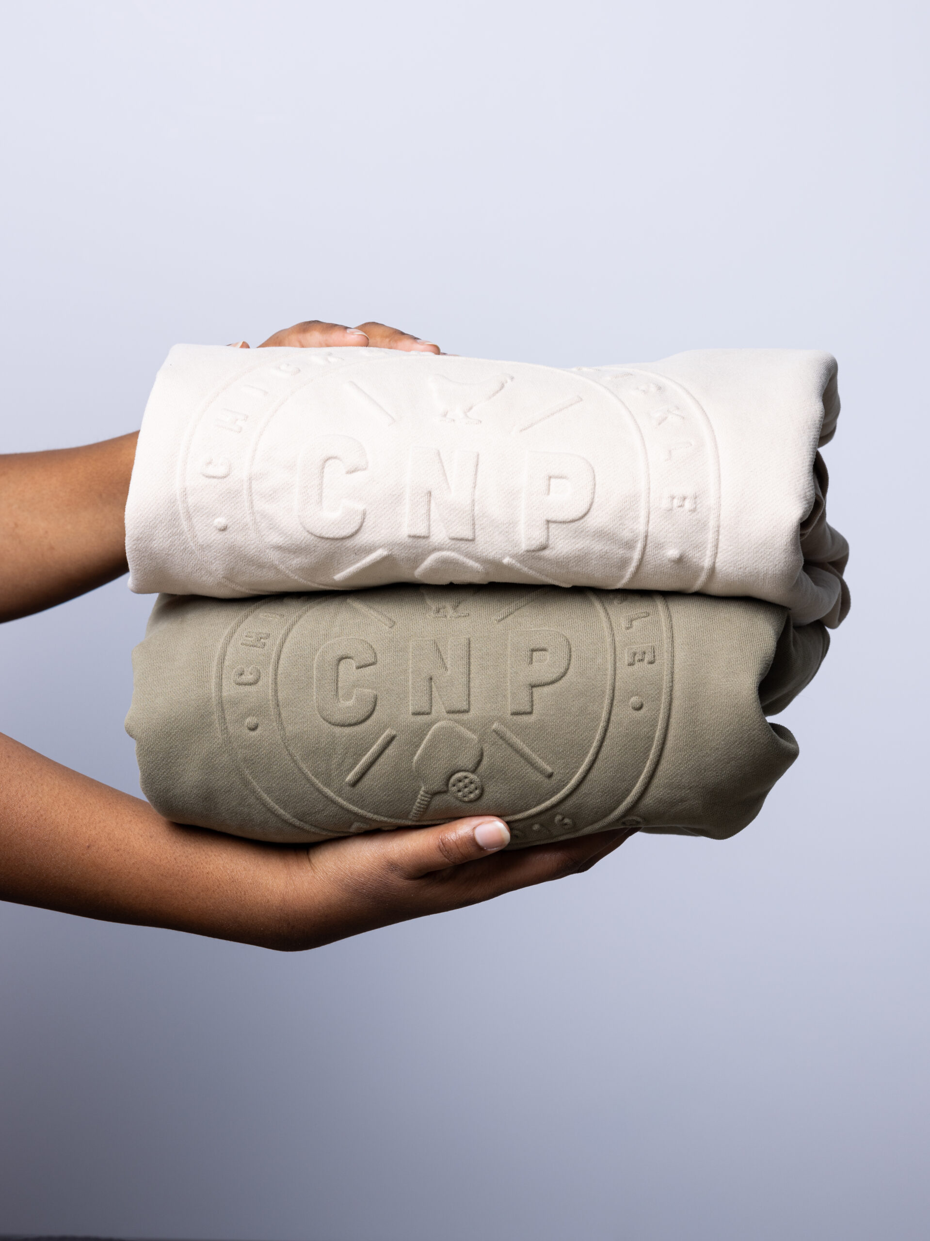 A person holds two folded sweatshirts, one beige and one olive green, both featuring an embossed circular CNP logo design on the front against a plain light background.