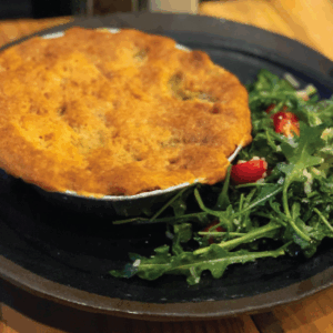 A golden-brown pot pie sits on a dark plate next to a side of fresh arugula salad with cherry tomatoes. The dish is placed on a wooden table.