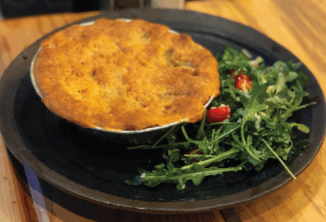 A golden-brown pot pie sits on a dark plate next to a side of fresh arugula salad with cherry tomatoes. The dish is placed on a wooden table.