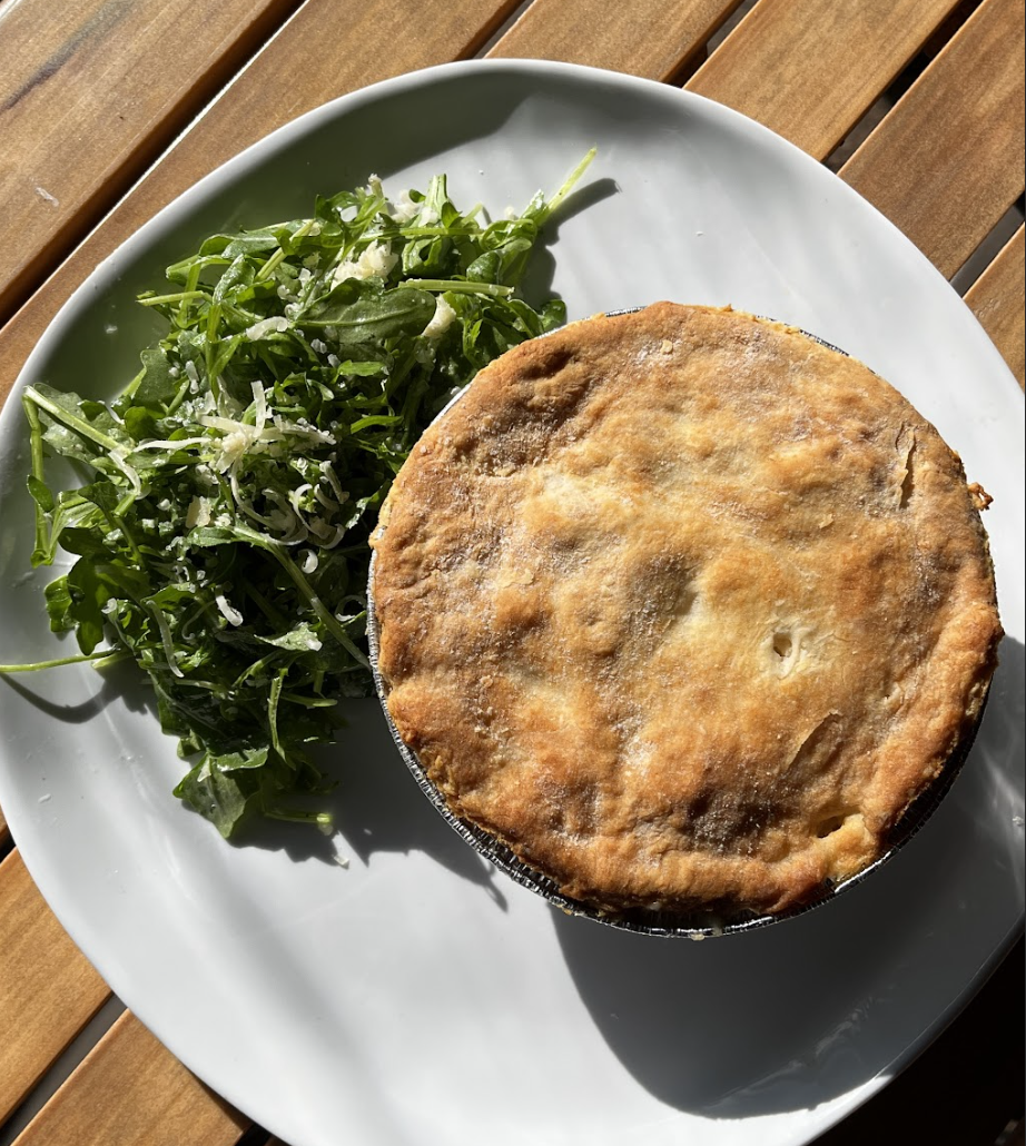 A golden-brown savory pot pie sits on a white plate next to a fresh green arugula salad with grated cheese, just like you'd find on the Chicken N Picklle Menu, all arranged on a wooden table in sunlight.