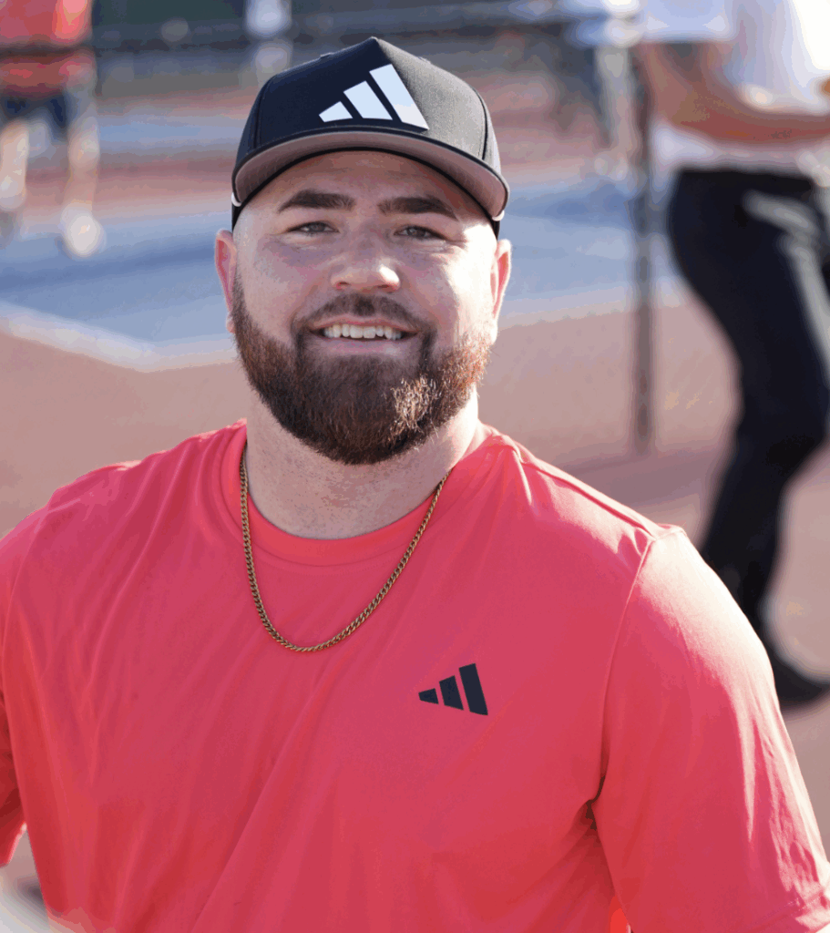 A bearded man wearing a black adidas cap, a gold chain, and a bright coral adidas shirt smiles outdoors near a tennis or pickleball court, with other people and a fence in the background.