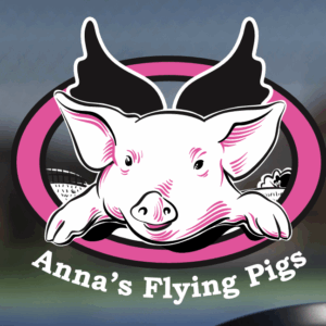 Cartoon illustration of a smiling pig with black wings inside a pink and black oval, with the text Anna’s Flying Pigs in white below the image.