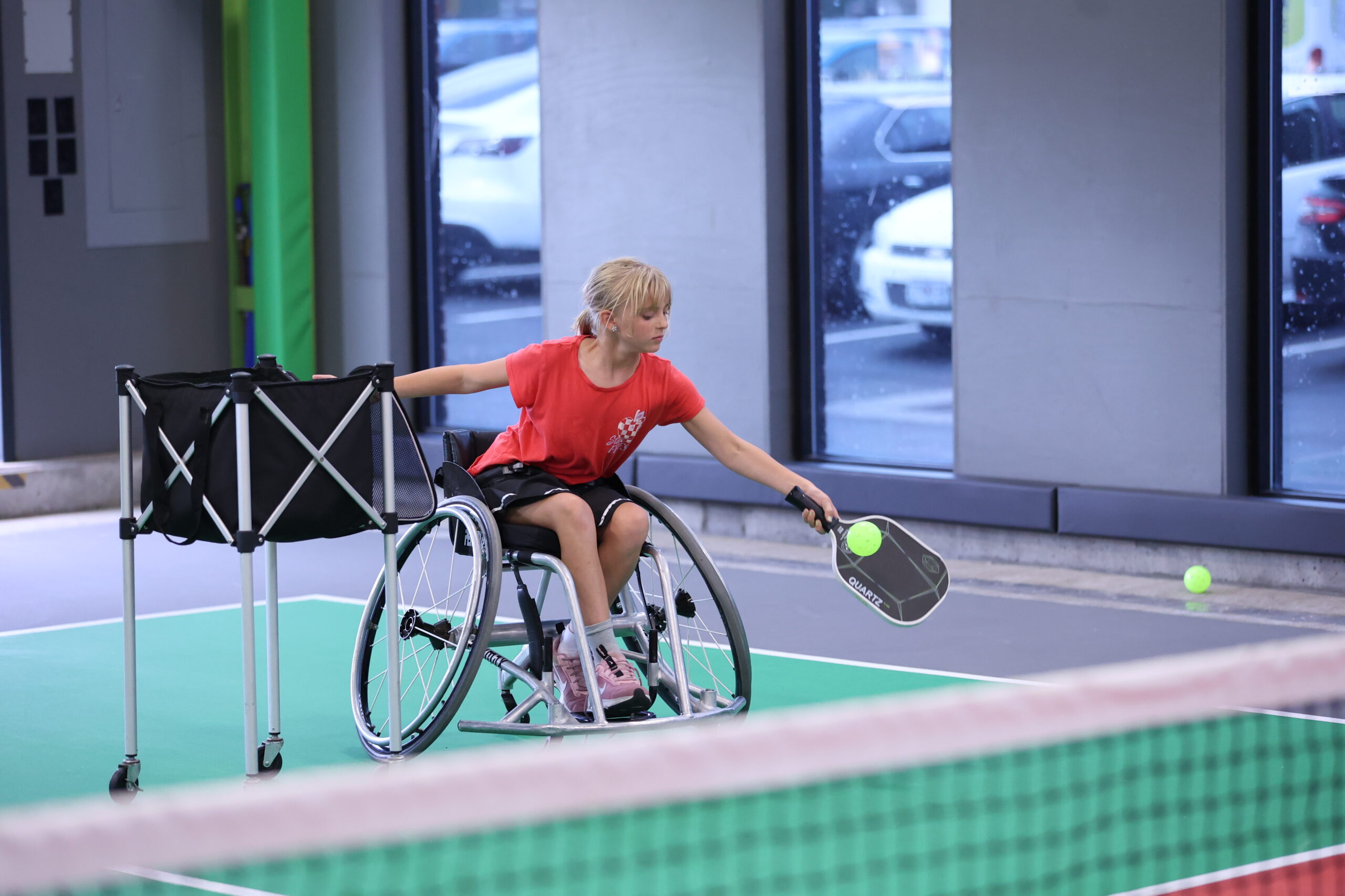 A young girl in a wheelchair plays pickleball indoors, reaching out with a paddle to hit a ball. She is on a green court with a net and a cart of balls nearby. Cars are visible outside the large windows in the background.