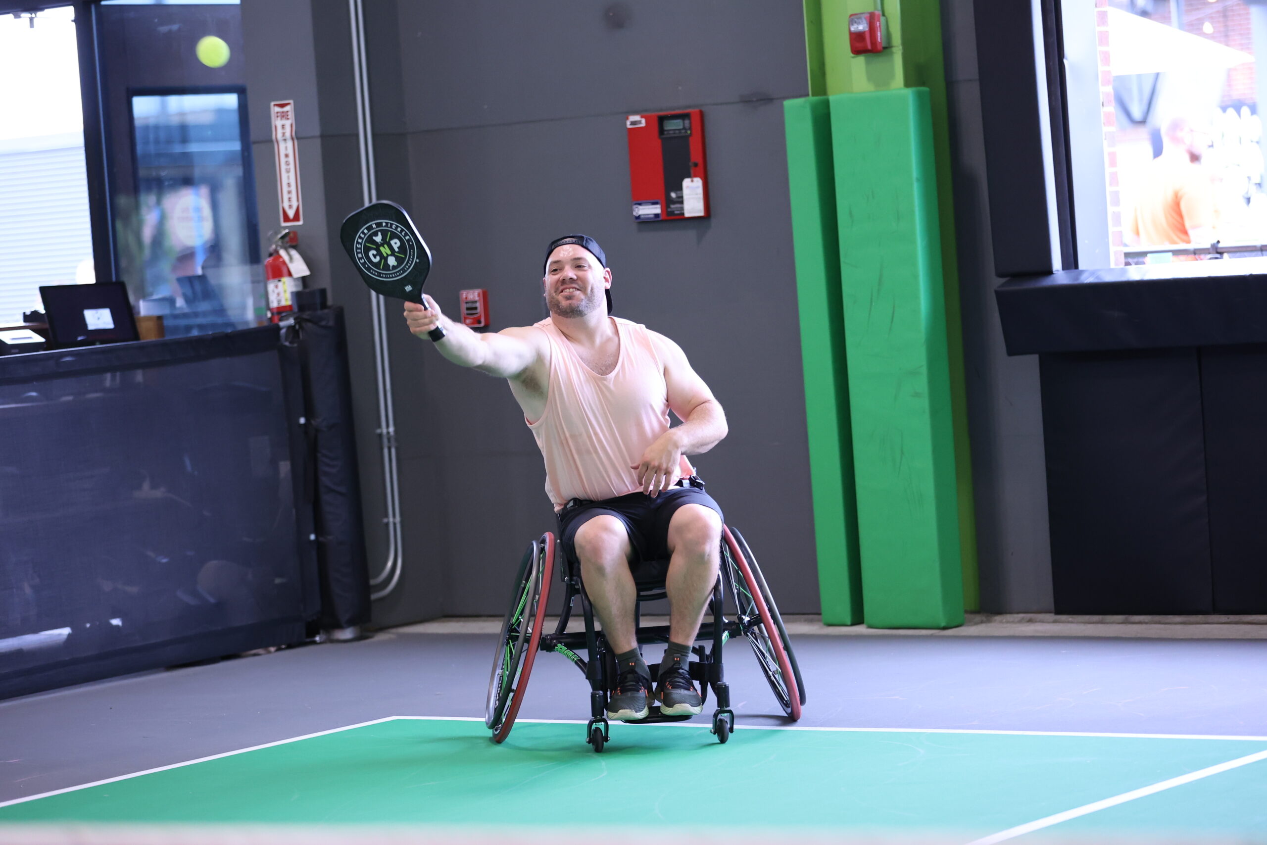 A man in a wheelchair plays pickleball indoors, wearing a light tank top, shorts, and a cap, reaching out with a paddle to hit the ball.