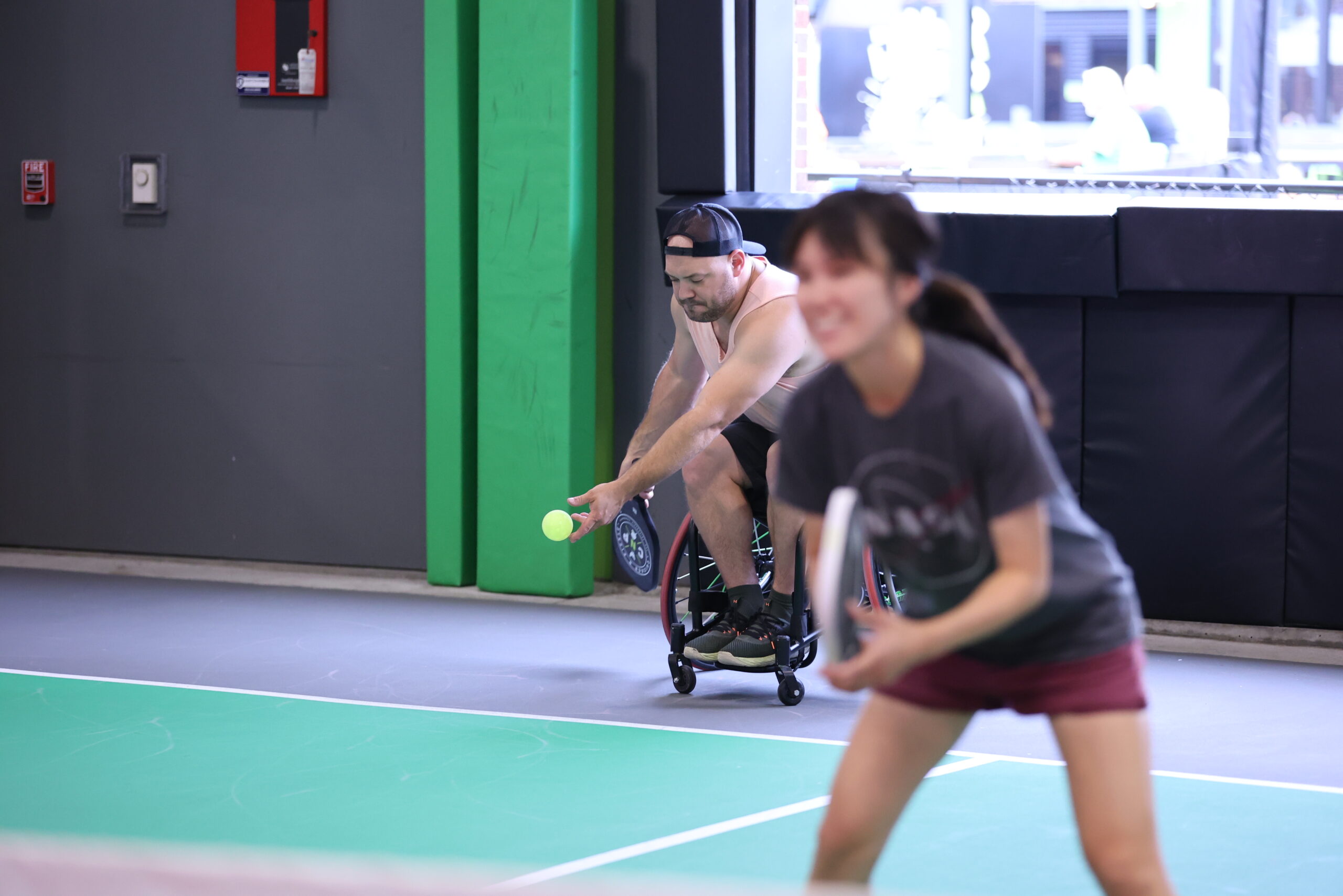 A man in a wheelchair prepares to hit a pickleball on an indoor court while a woman in the foreground stands ready, holding a paddle and smiling.