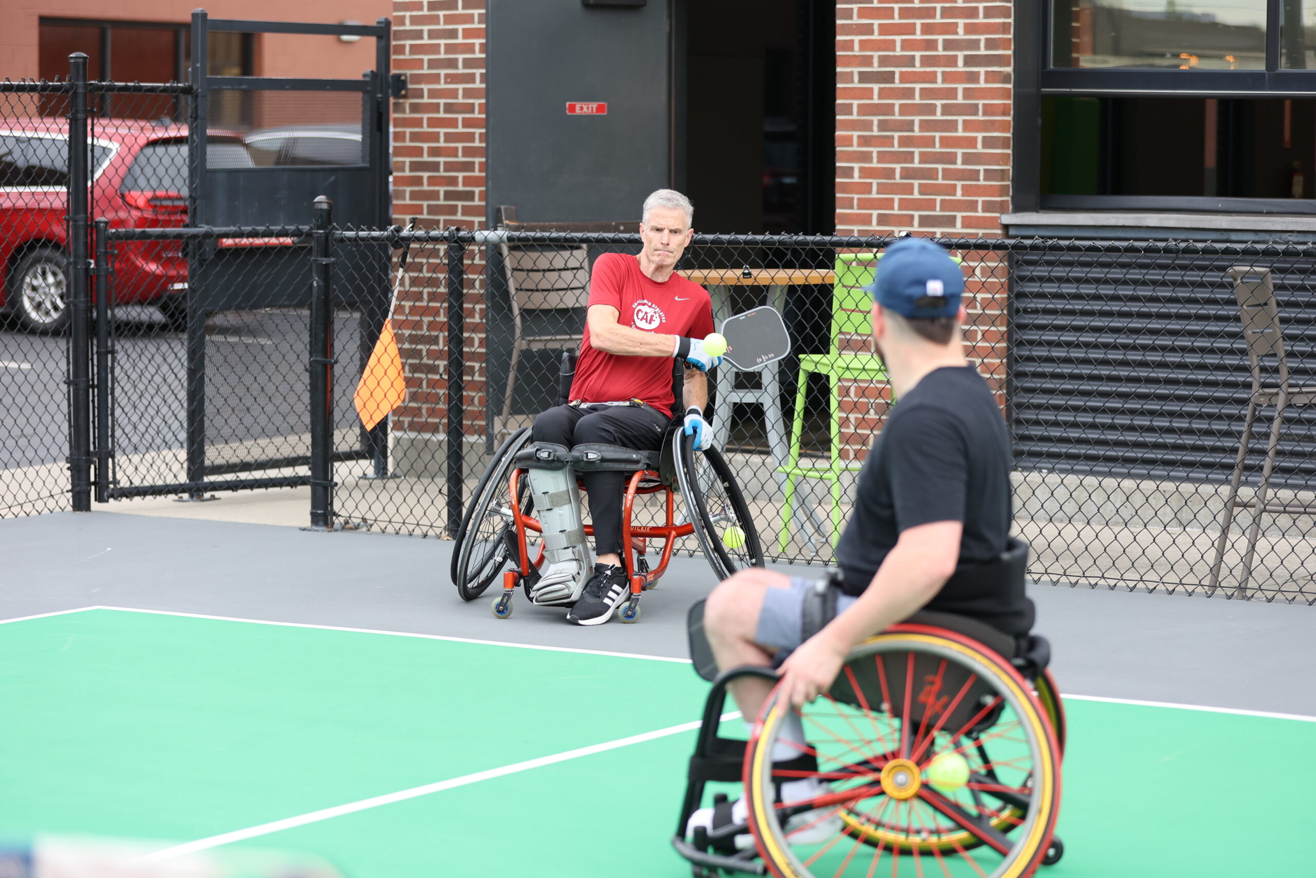 Two men in wheelchairs play a sport on an outdoor court; one man in a red shirt holds a racket while sitting near a fence, and another man in a black shirt faces him in the foreground.