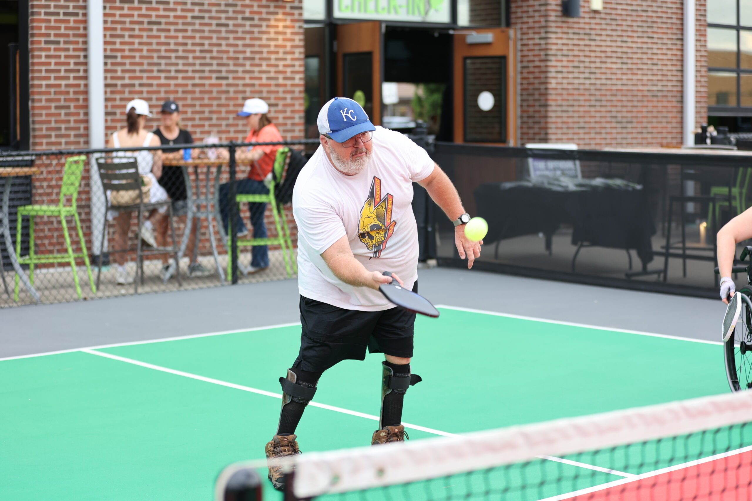 An older man wearing a cap and knee braces plays pickleball, reaching to hit a ball with his paddle on an outdoor court. People sit at tables behind a fence in the background.