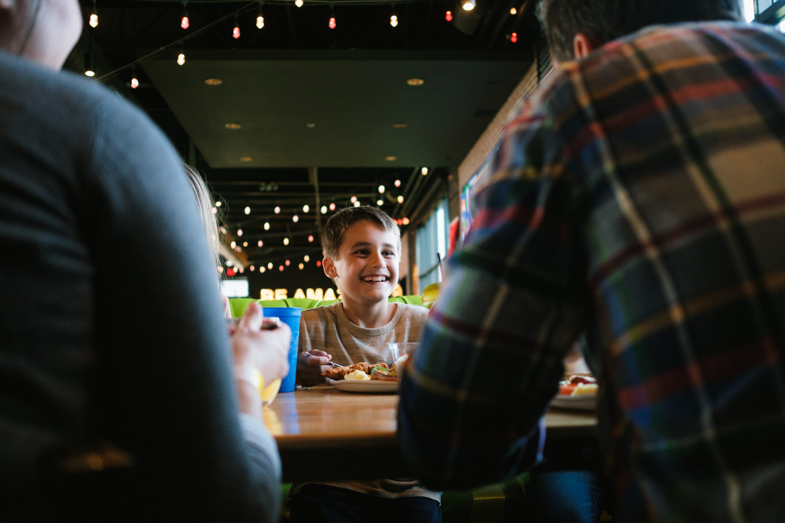 A smiling child sits at a restaurant table with two adults, who are seen from behind. String lights hang from the ceiling, creating a warm and inviting atmosphere.