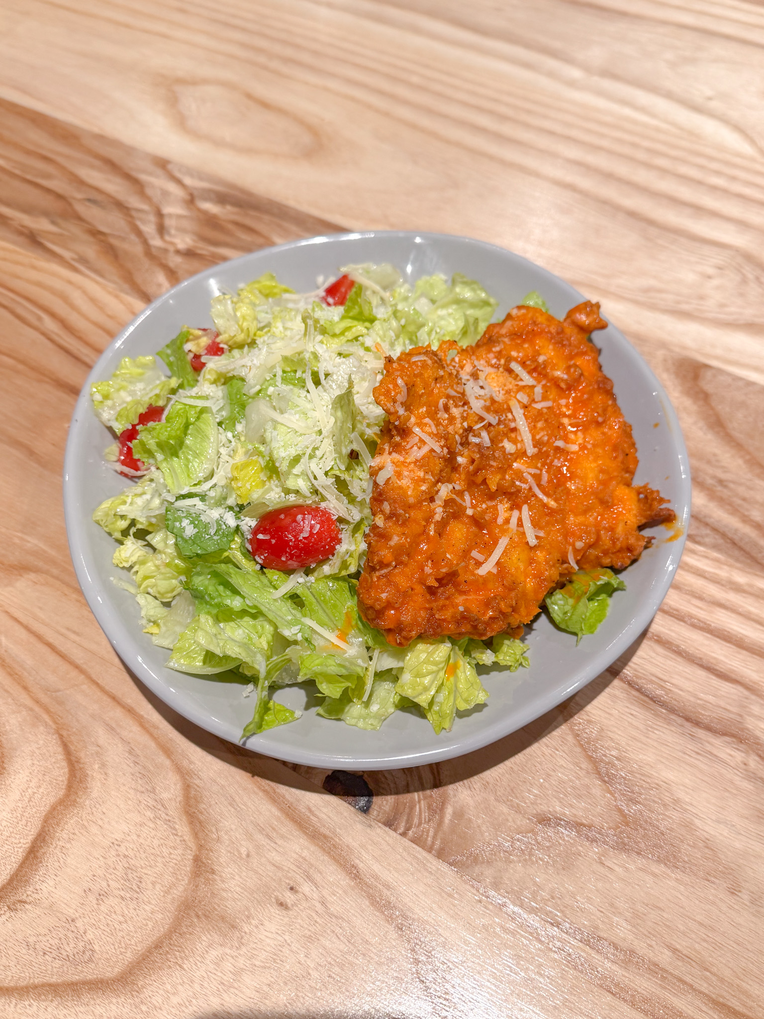 A plate with Caesar salad, topped with shredded cheese and cherry tomatoes, next to a piece of breaded, sauced fried chicken on a light wooden table.