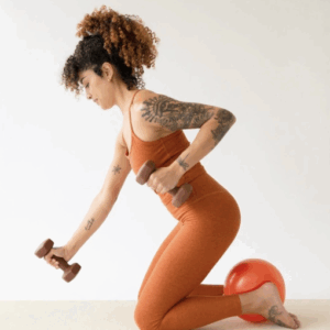 A woman in an orange workout outfit kneels on the floor with a red exercise ball under her shin, holding two dumbbells and performing a row exercise. She has curly hair and visible arm tattoos.