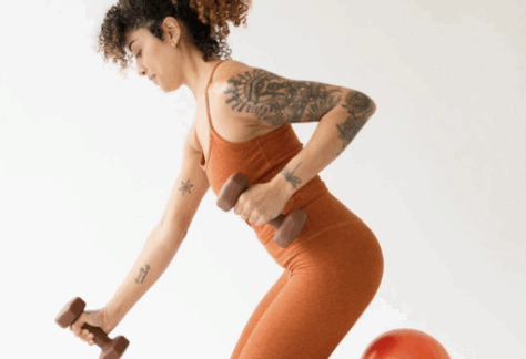 A woman in an orange workout outfit kneels on the floor with a red exercise ball under her shin, holding two dumbbells and performing a row exercise. She has curly hair and visible arm tattoos.