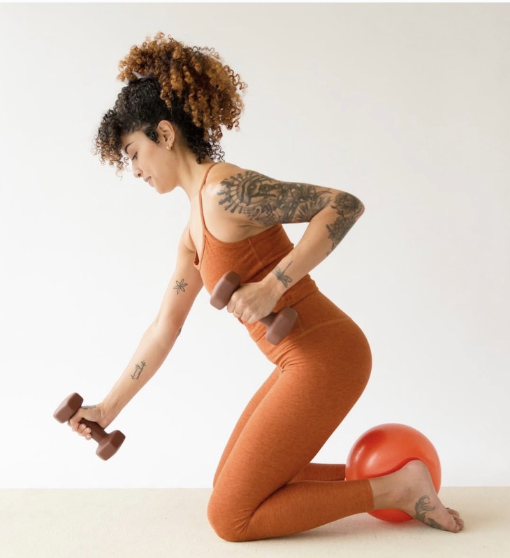A woman in an orange workout outfit kneels on the floor with a red exercise ball under her shin, holding two dumbbells and performing a row exercise. She has curly hair and visible arm tattoos.