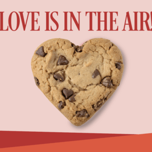A heart-shaped chocolate chip cookie sits below the bold red text LOVE IS IN THE AIR! on a pink background with red accents at the bottom.