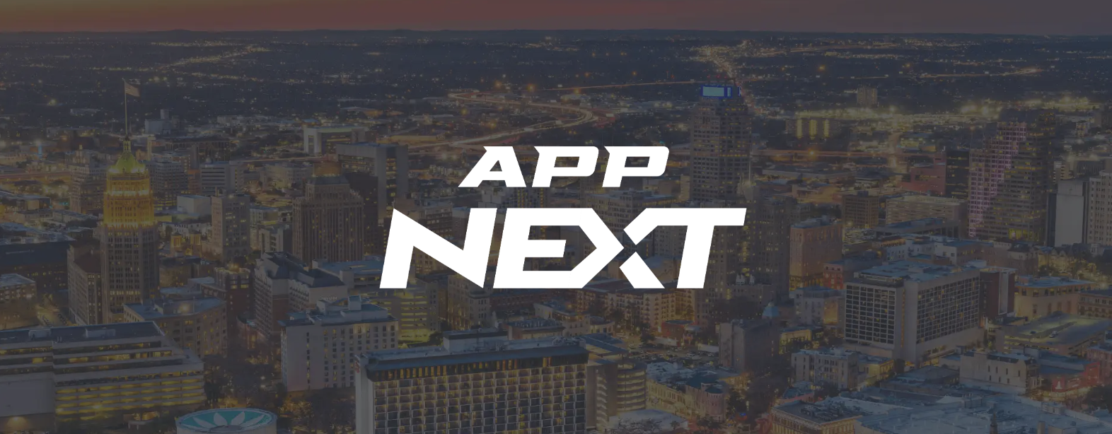 City skyline at dusk with illuminated buildings and streets. The bold white text APP NEXT is centered over the image, partially obscuring the city view in the background.