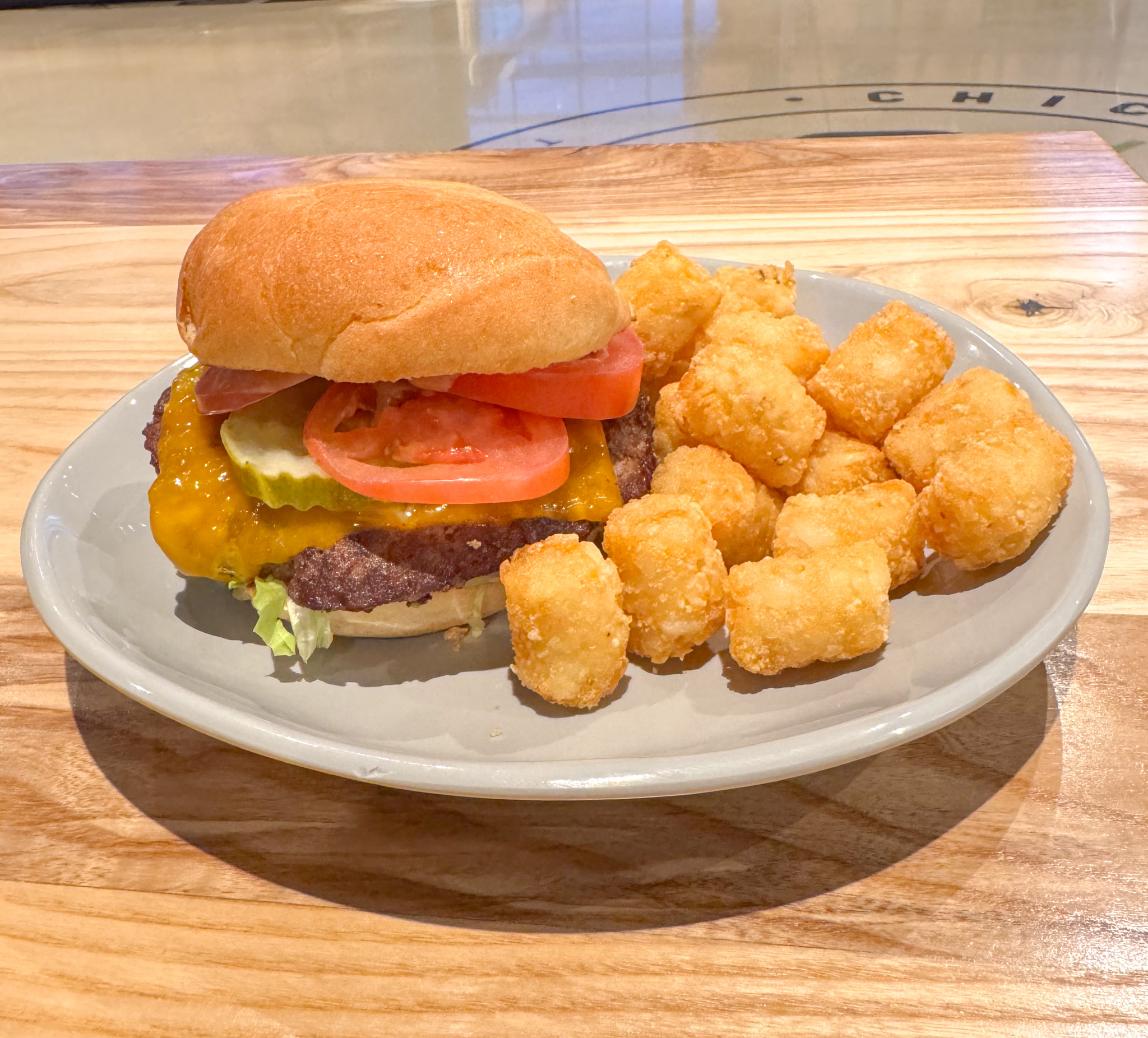 A cheeseburger with lettuce, tomato, pickles, and cheddar cheese is served on a plate next to a portion of crispy tater tots, all placed on a wooden table.