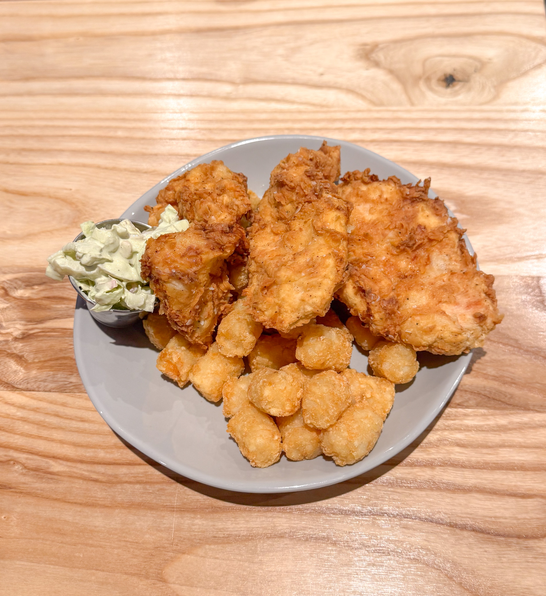 A gray plate with golden-brown fried chicken, crispy tater tots, and a small serving of coleslaw sits on a wooden table.