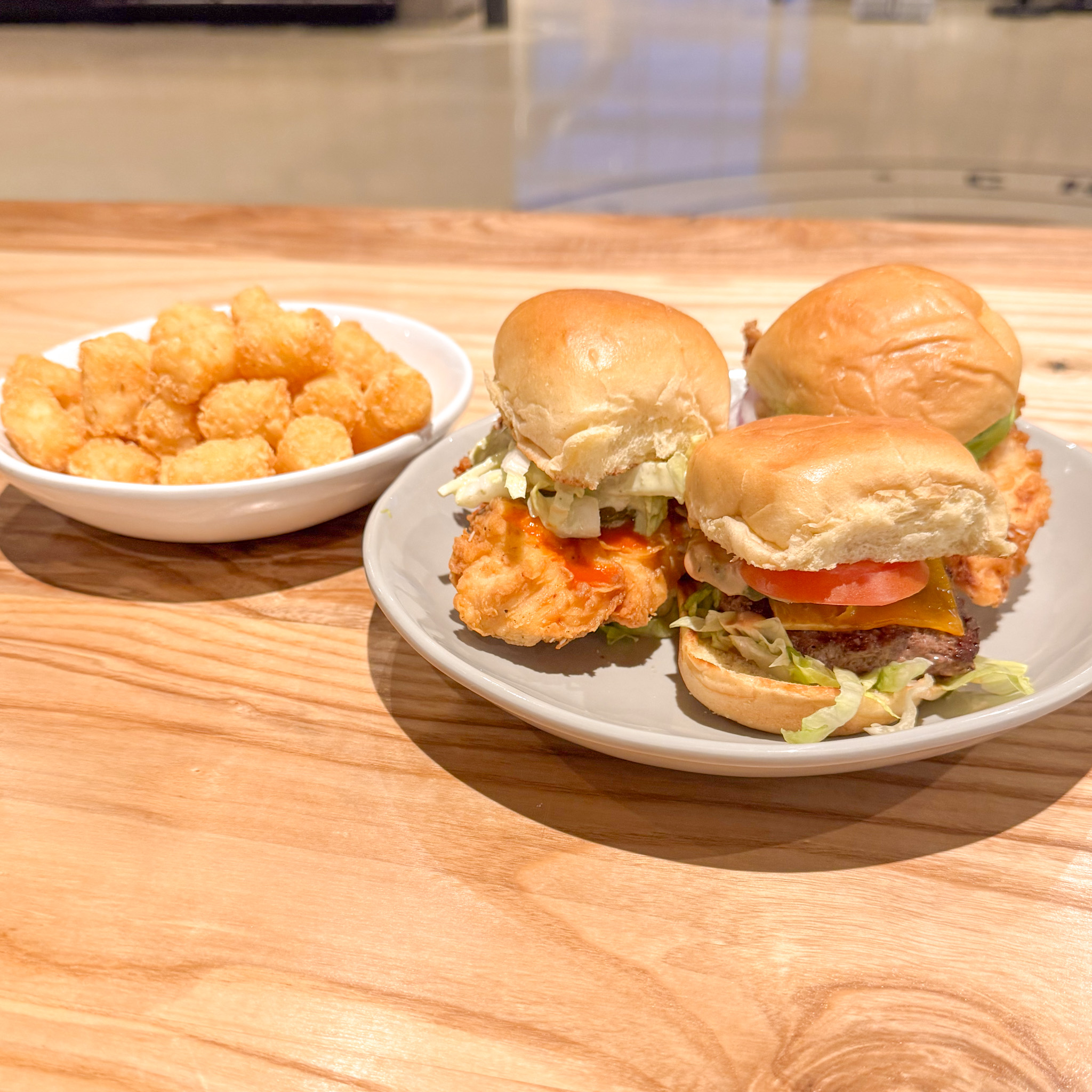 A plate with three sliders, including fried chicken and beef patties with lettuce and cheese, sits next to a bowl of crispy tater tots on a wooden table.