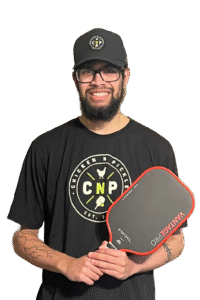 A smiling man with glasses, a beard, and a black cap wears a Chicken N Pickle t-shirt and holds a black and red pickleball paddle. He has tattoos on his right forearm. The background is plain white.