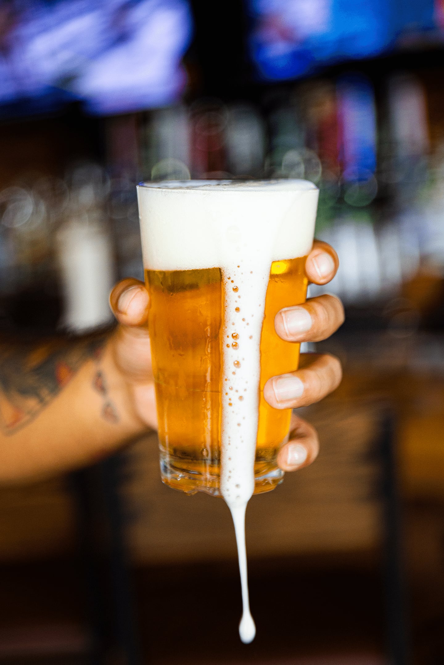 A hand holds a glass of beer with a thick foamy head spilling over the side and dripping down, against a blurred bar background.