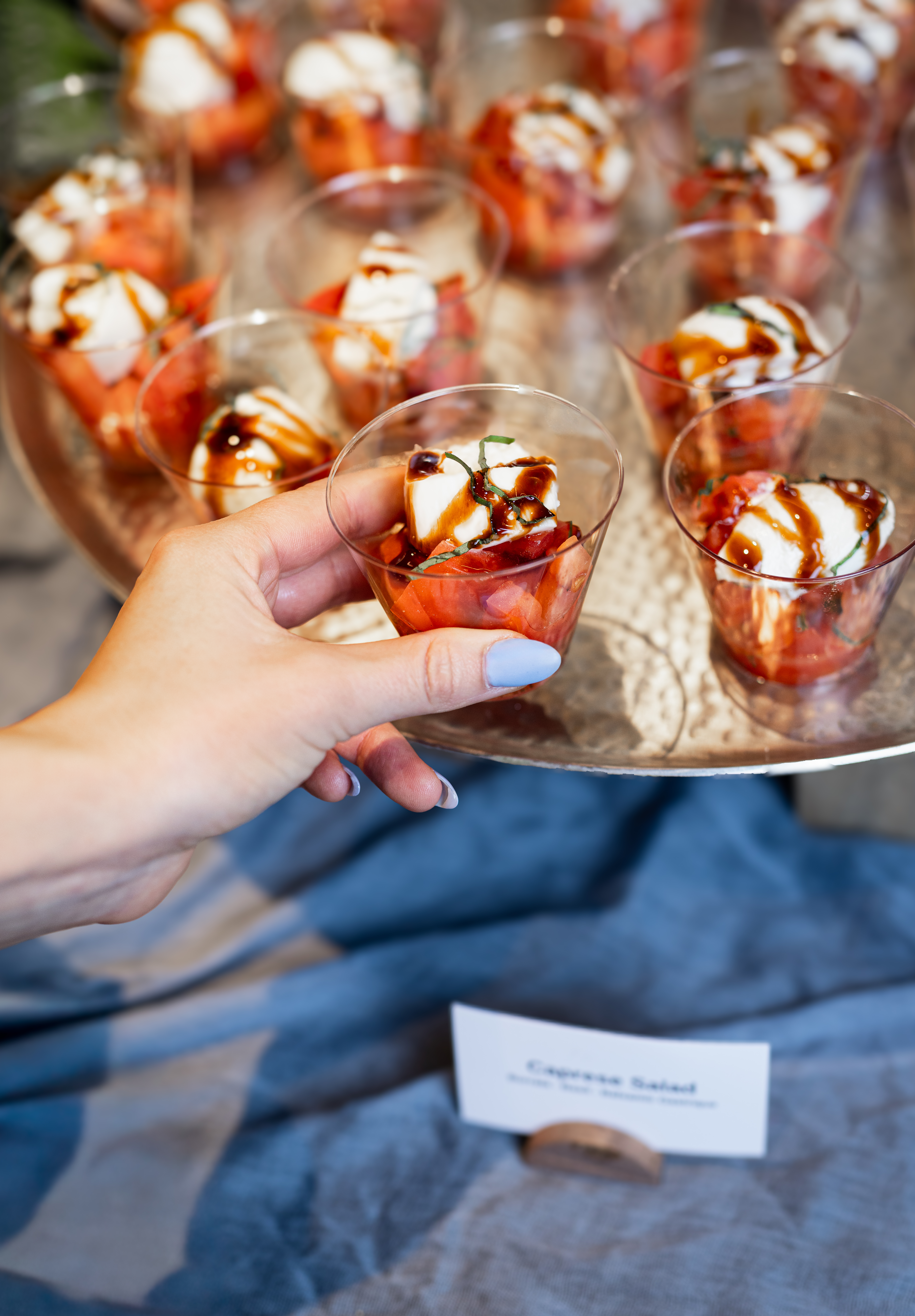 A hand picks up a small plastic cup filled with caprese salad, topped with mozzarella, tomato, basil, and balsamic glaze. Several other cups are arranged on a silver tray, with a label reading Caprese Salad.
