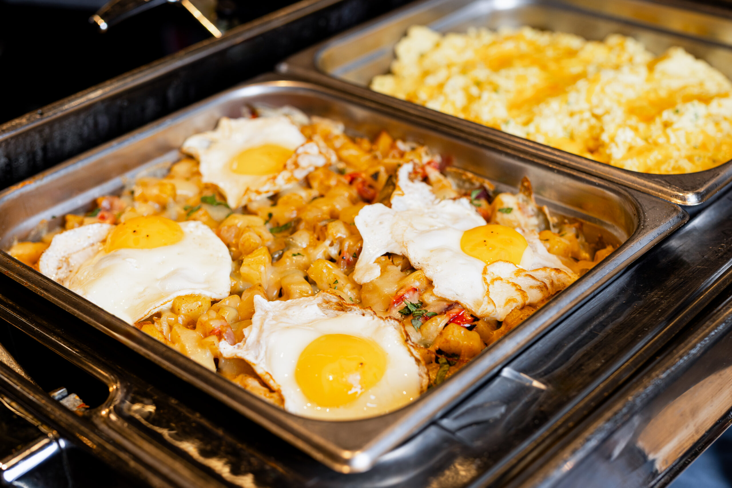 A metal tray filled with a breakfast hash topped with sunny-side-up eggs sits next to a tray of scrambled eggs in a buffet setting.