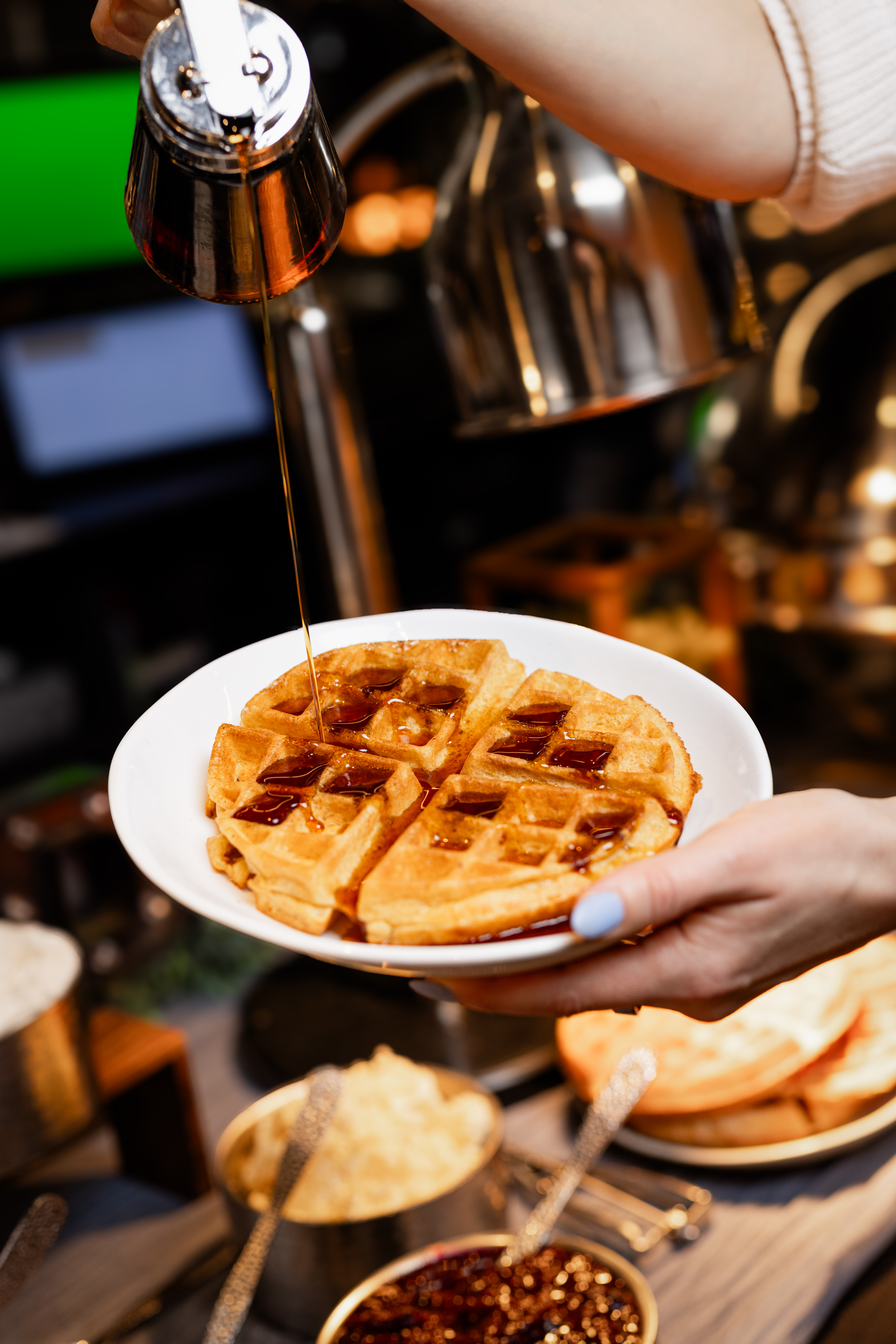 A hand holds a plate with a golden waffle, while syrup is being poured over it. The background shows various food items and utensils in a warm, inviting setting.