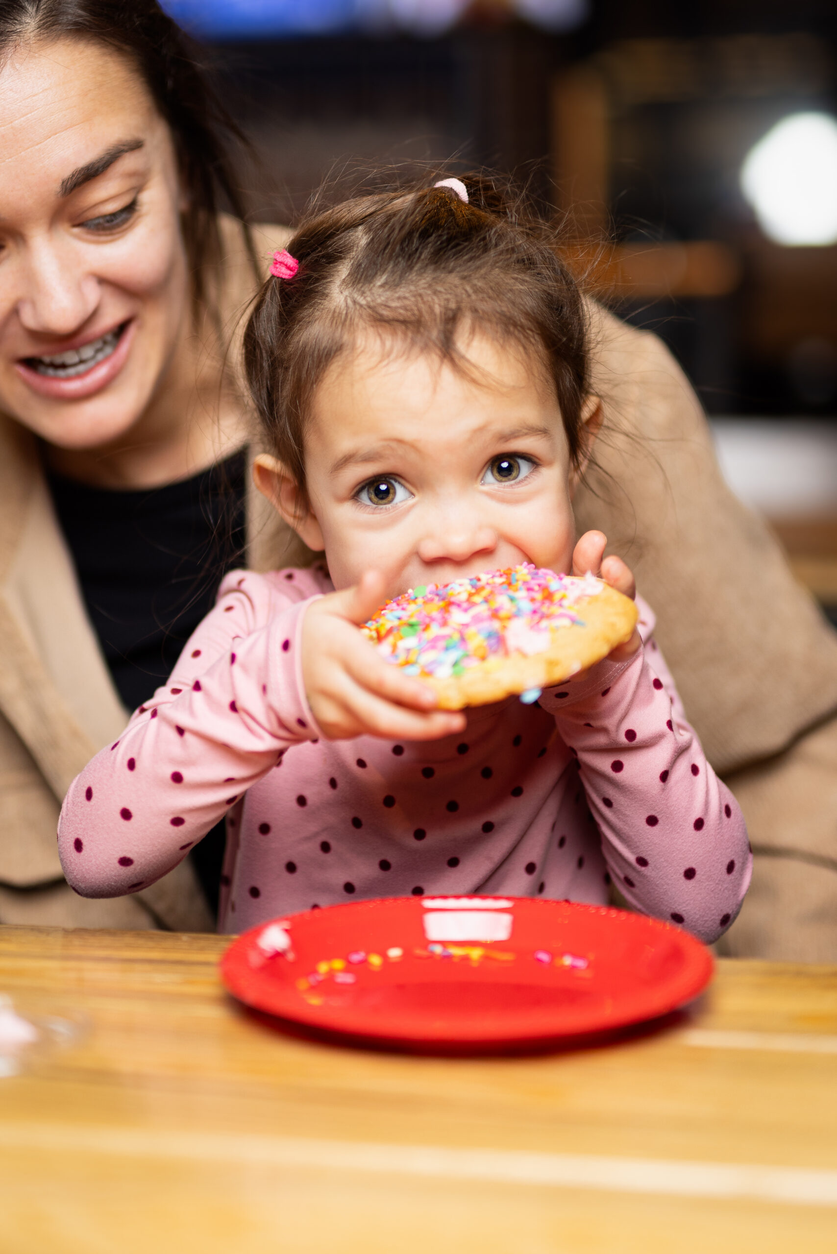 A young girl with brown hair in pigtails is about to bite into a large, sprinkle-covered cookie, while a smiling woman sits beside her. They are seated at a wooden table with a red plate.