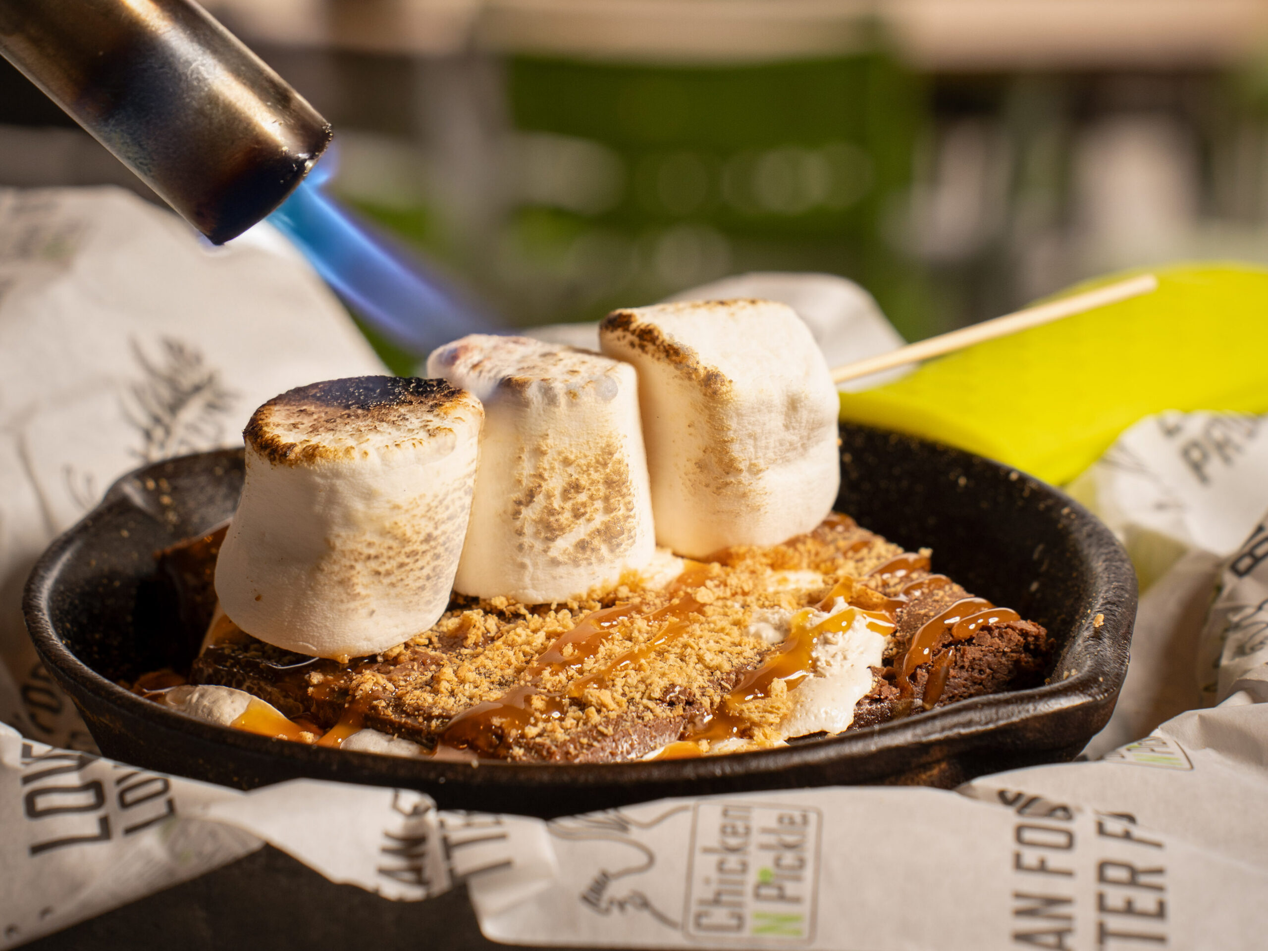 A torch browns four marshmallows on top of a dessert in a small black skillet, set on paper-lined dish. Outdoor seating and green chairs appear blurred in the background.