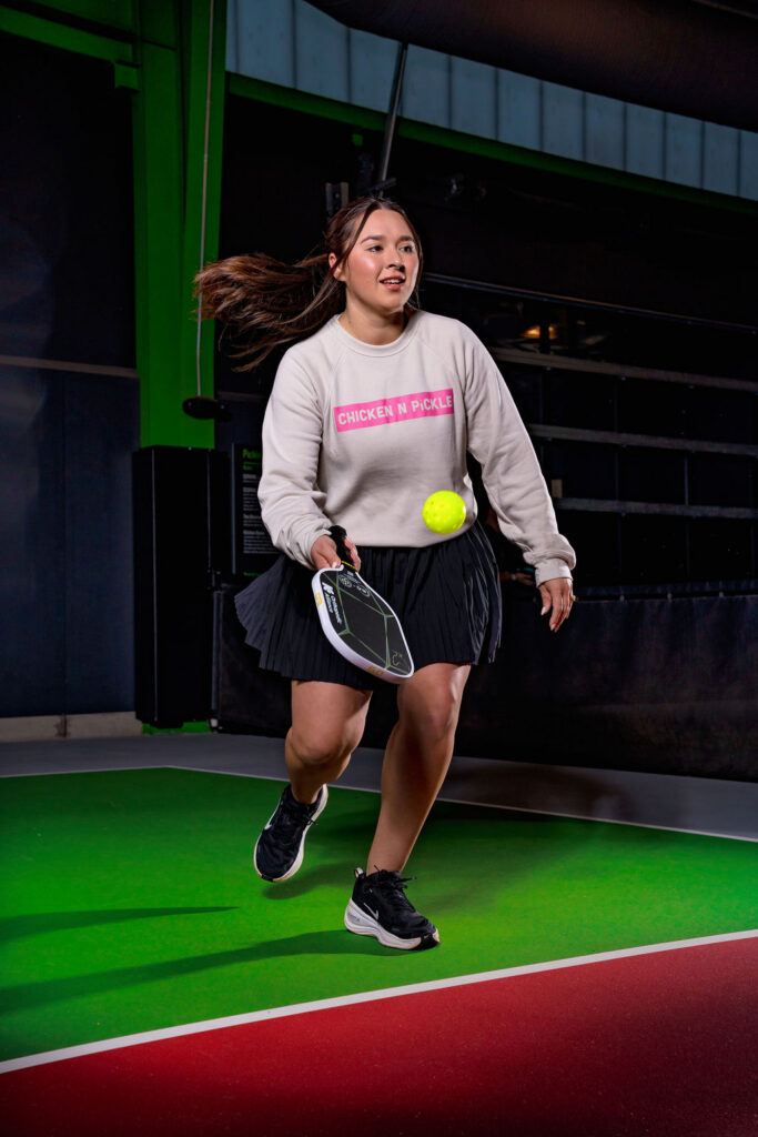 A woman playing pickleball indoors, wearing a CHICKEN N PICKLE sweatshirt, black skirt, and sneakers, about to hit a yellow ball with her paddle on a green and red court.
