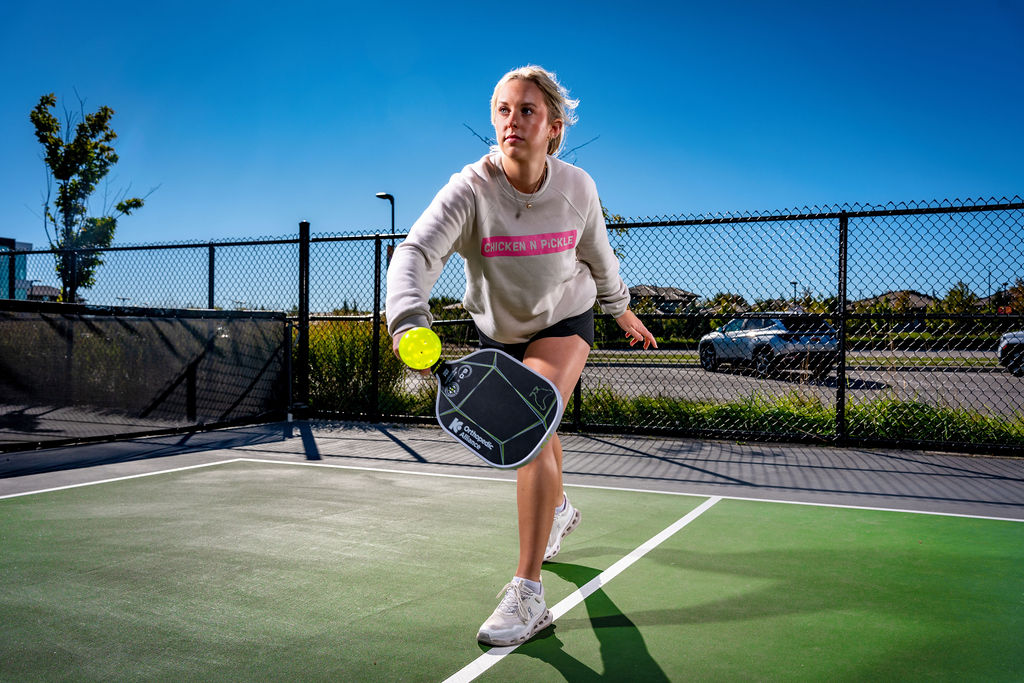 A woman plays pickleball on an outdoor court, wearing a white sweatshirt and sneakers, focused on hitting the ball with her paddle under a clear blue sky.
