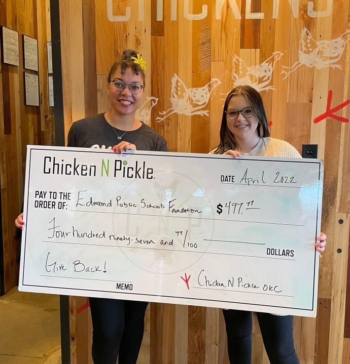 Two smiling women hold a large check from Chicken N Pickle for $497.77, made out to the Edmond Public Schools Foundation, dated April 2022, with the memo Give Back! written on it.