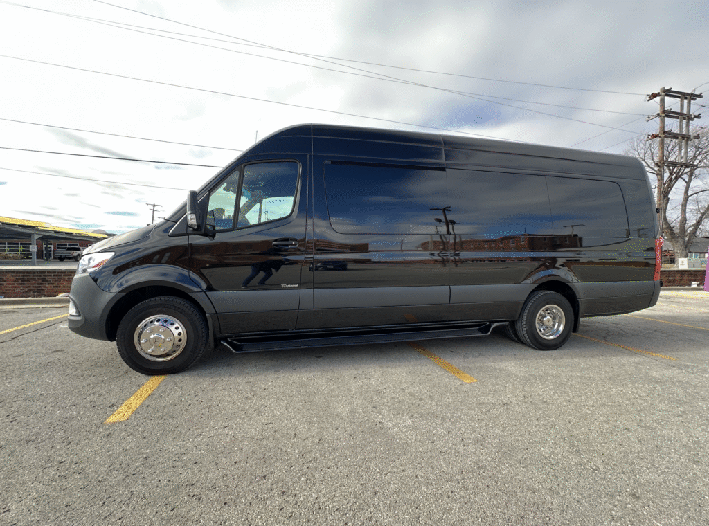 A black van is parked across two parking spaces in an outdoor lot on a cloudy day, with buildings and utility poles visible in the background.