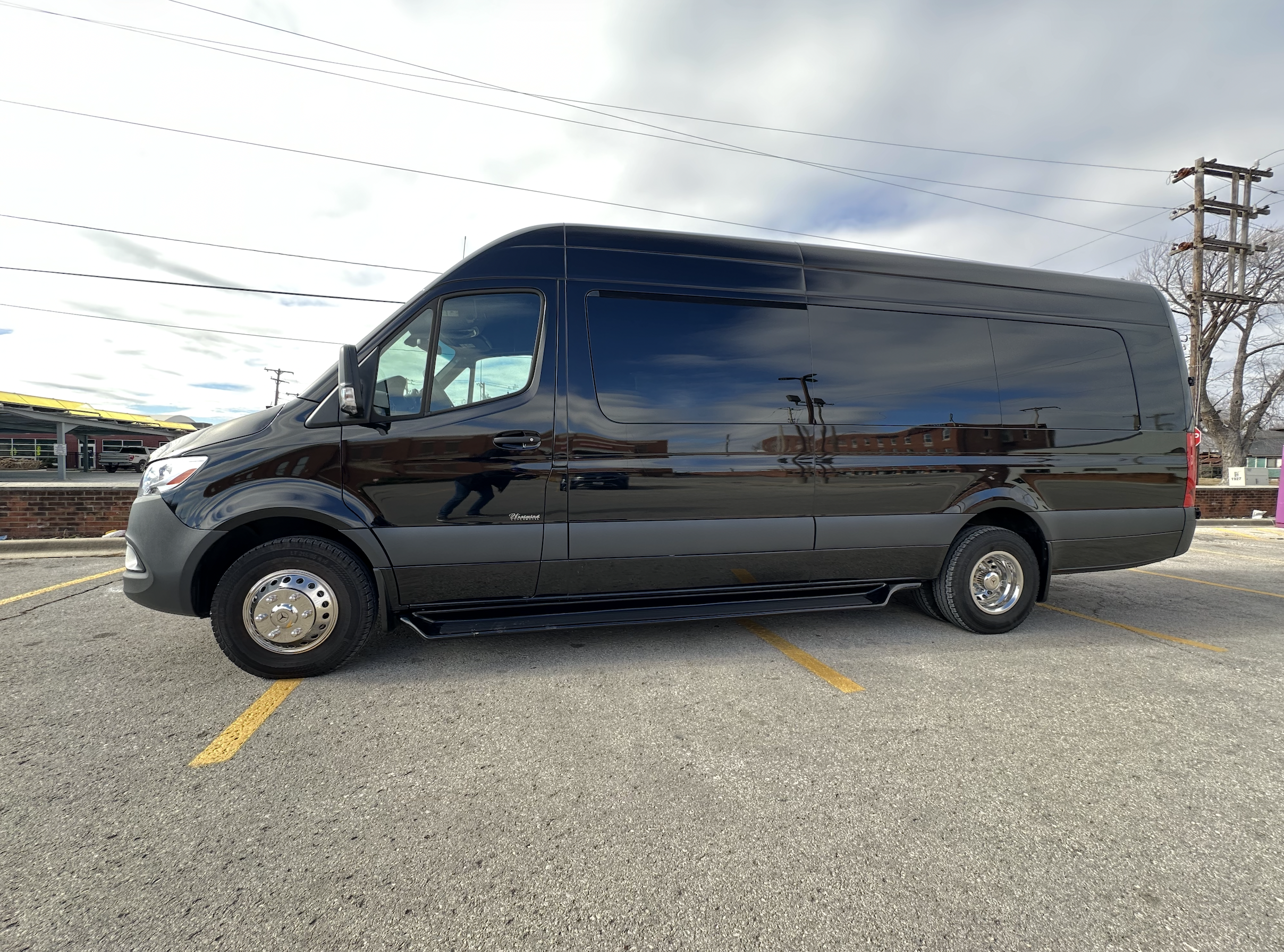 A black van is parked across two parking spaces in an outdoor lot on a cloudy day, with buildings and utility poles visible in the background.