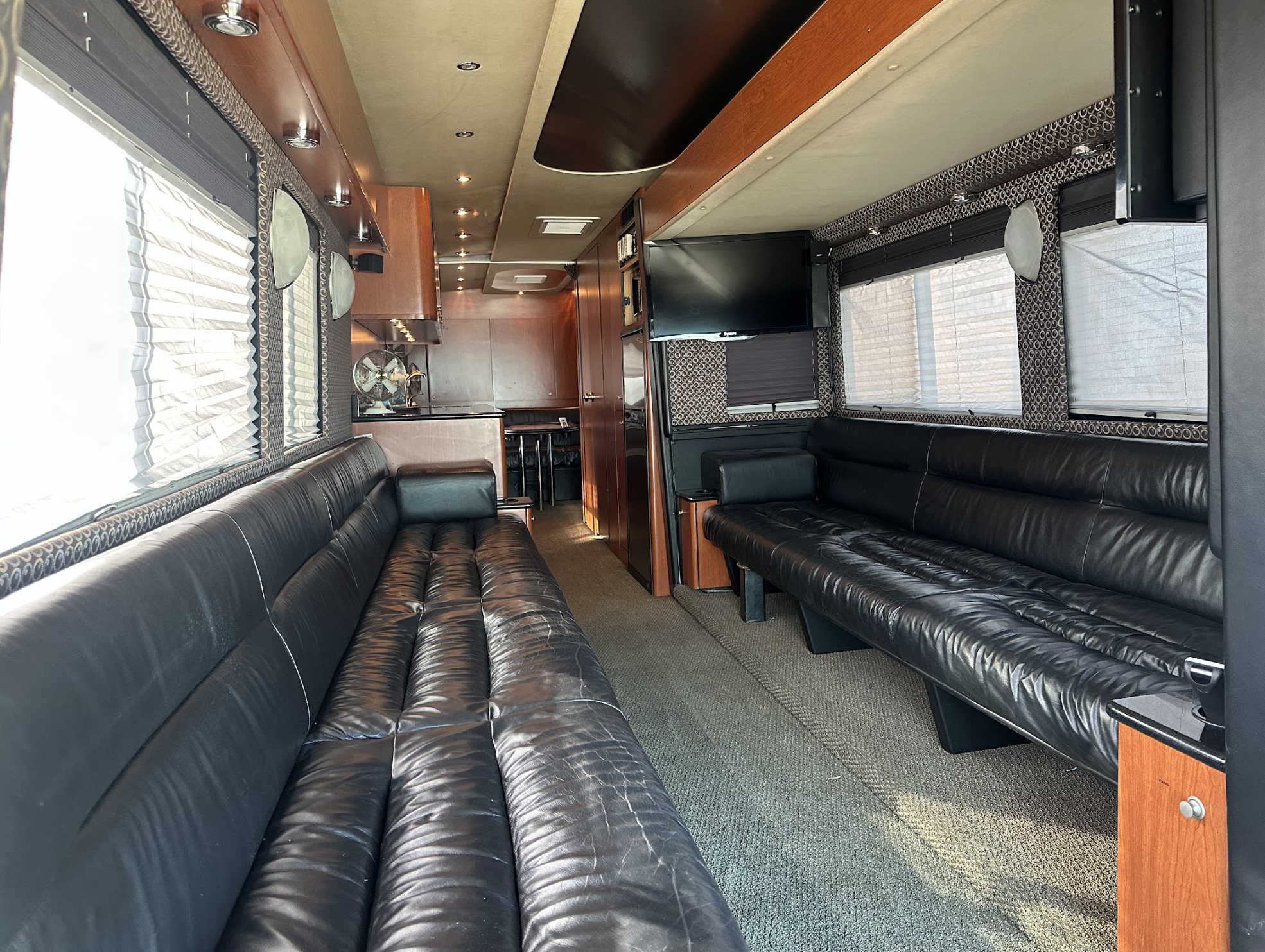 The interior of a modern tour bus with two long black leather couches on either side, large windows with blinds, a mounted TV, and a view toward a dining area and kitchenette in the back.