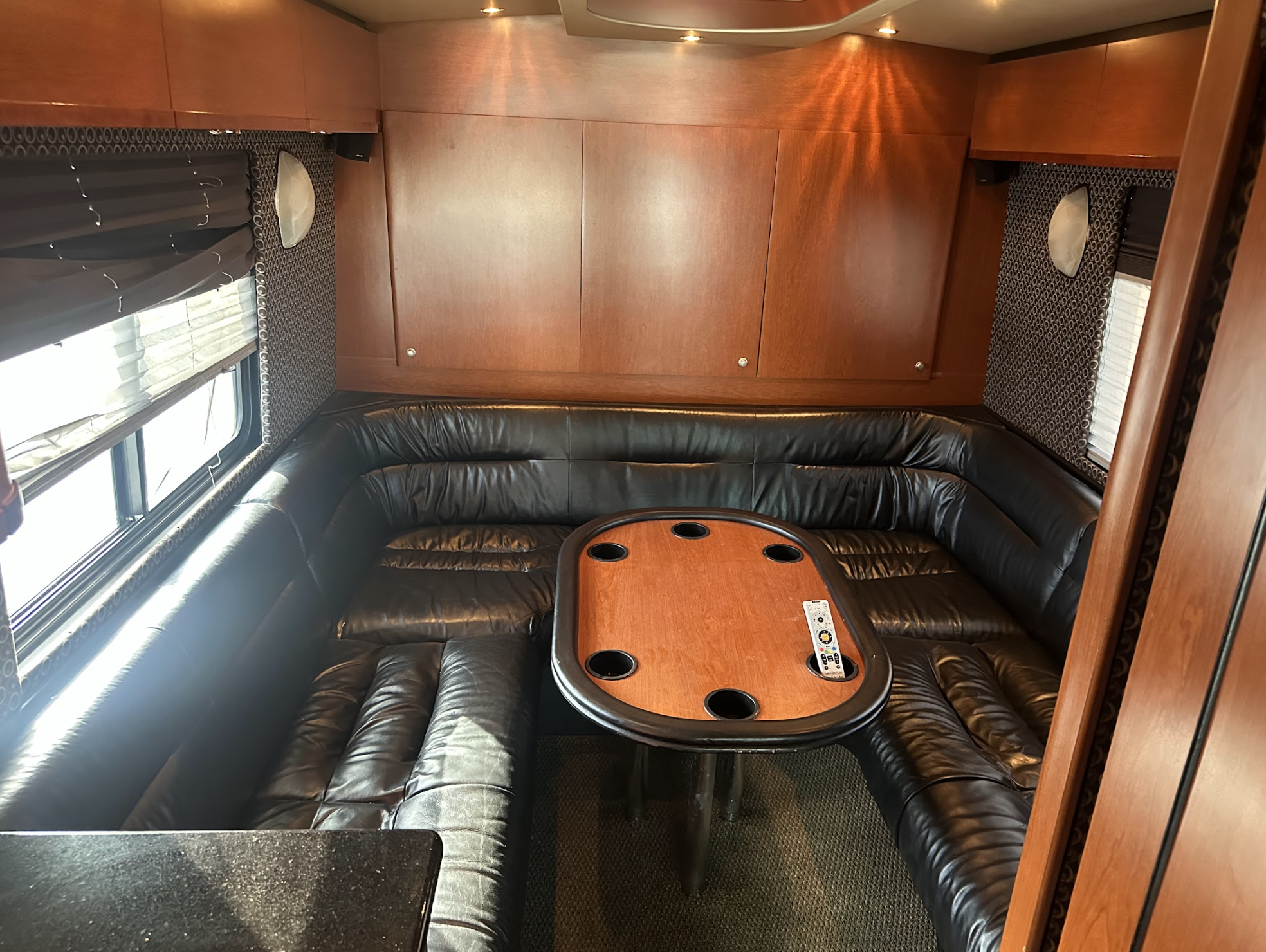 A cozy lounge area with black leather U-shaped seating around a wooden table with cup holders and a remote control, surrounded by wood-paneled walls and windows with blinds.