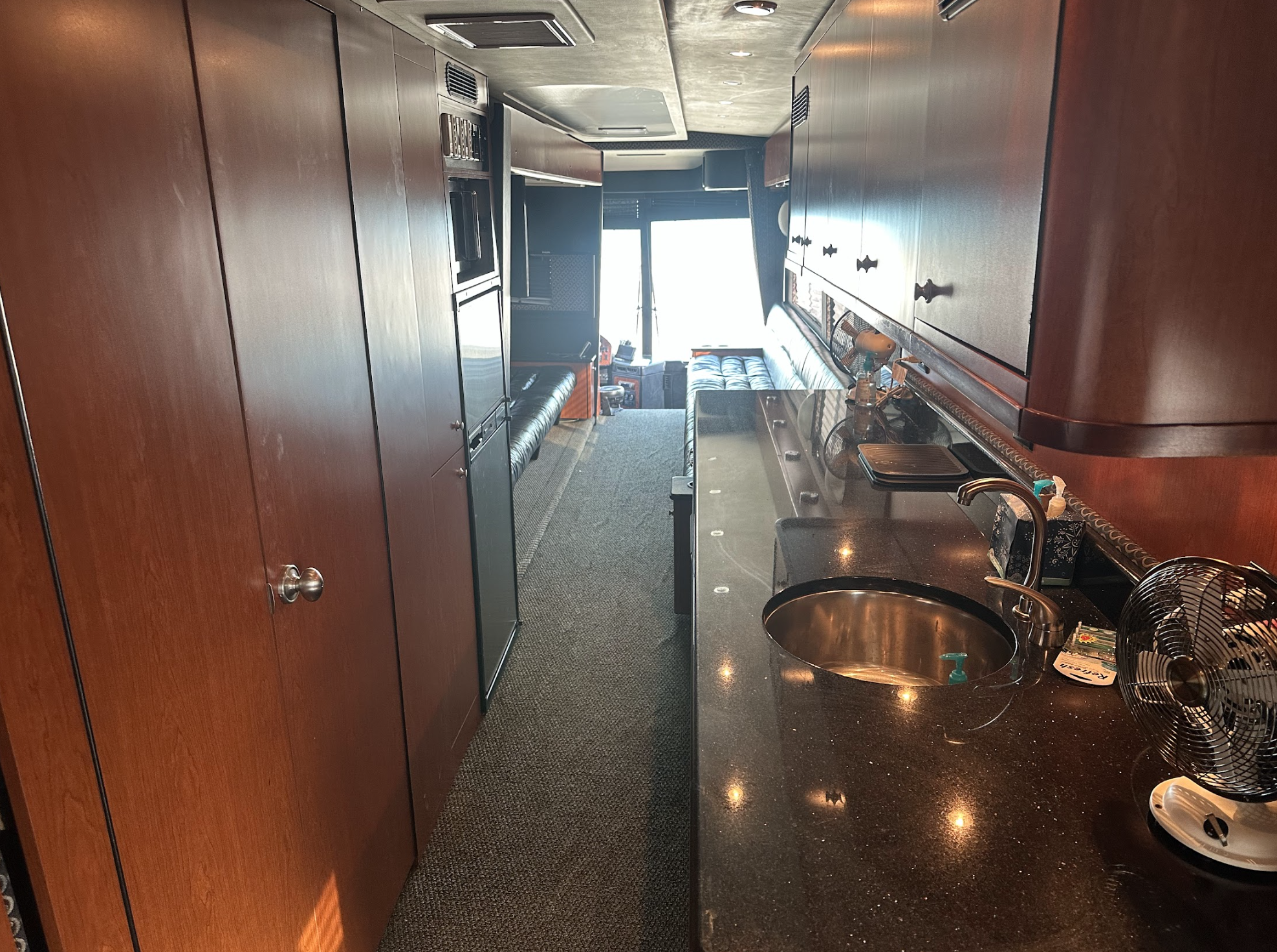 Interior of a motorhome or tour bus with wood cabinets, a black countertop with a sink, a small fan, seating area in the back, and natural light coming from windows at the far end.