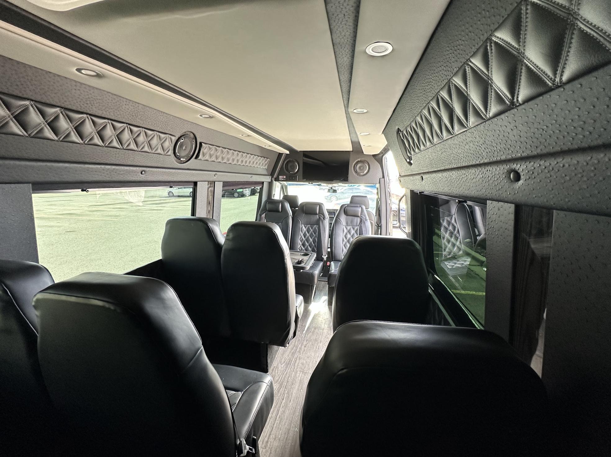 Interior of a luxury van with black leather seats, quilted wall panels, and large windows. Several seats face each other around a small table, and sunlight streams in from outside.