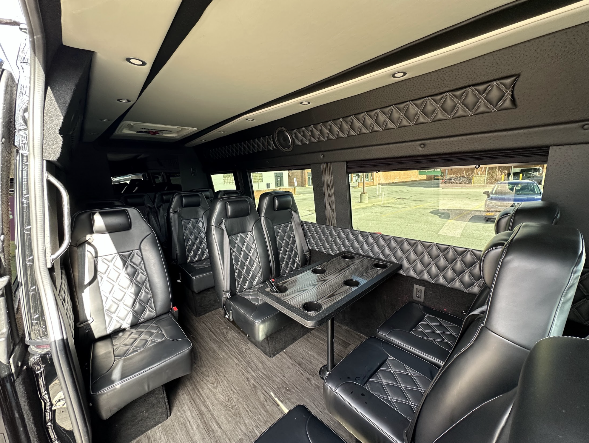 The image shows the interior of a luxury van with black leather seats, diamond-stitched upholstery, a table with cup holders, and large windows. The floor is wood-style, and the space looks clean and spacious.