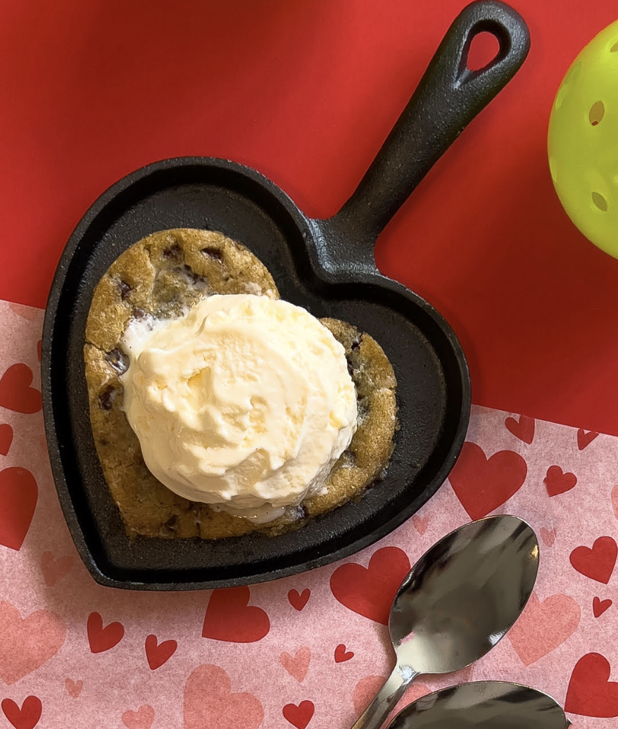 A heart-shaped skillet holds a chocolate chip cookie topped with vanilla ice cream, placed on red and pink heart-themed paper next to two spoons.