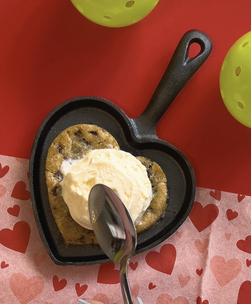 A heart-shaped skillet with a chocolate chip cookie topped with a scoop of vanilla ice cream. A spoon is about to scoop the dessert. The setup is on a red surface with a heart-patterned napkin and green objects nearby.