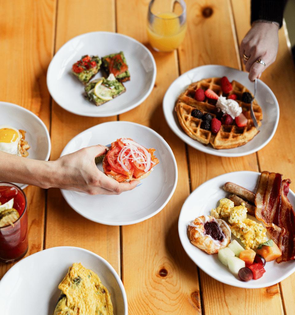 A table set with plates of breakfast foods including waffles with berries, avocado toast, an omelette, scrambled eggs with bacon and sausage, fruit, and a Bloody Mary. Two hands reach for food.