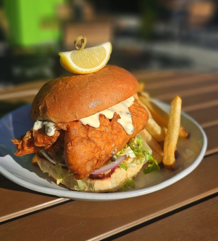 A crispy fried fish sandwich with lettuce, onions, and sauce in a bun, topped with a lemon wedge, served with French fries on a white plate on a wooden table.